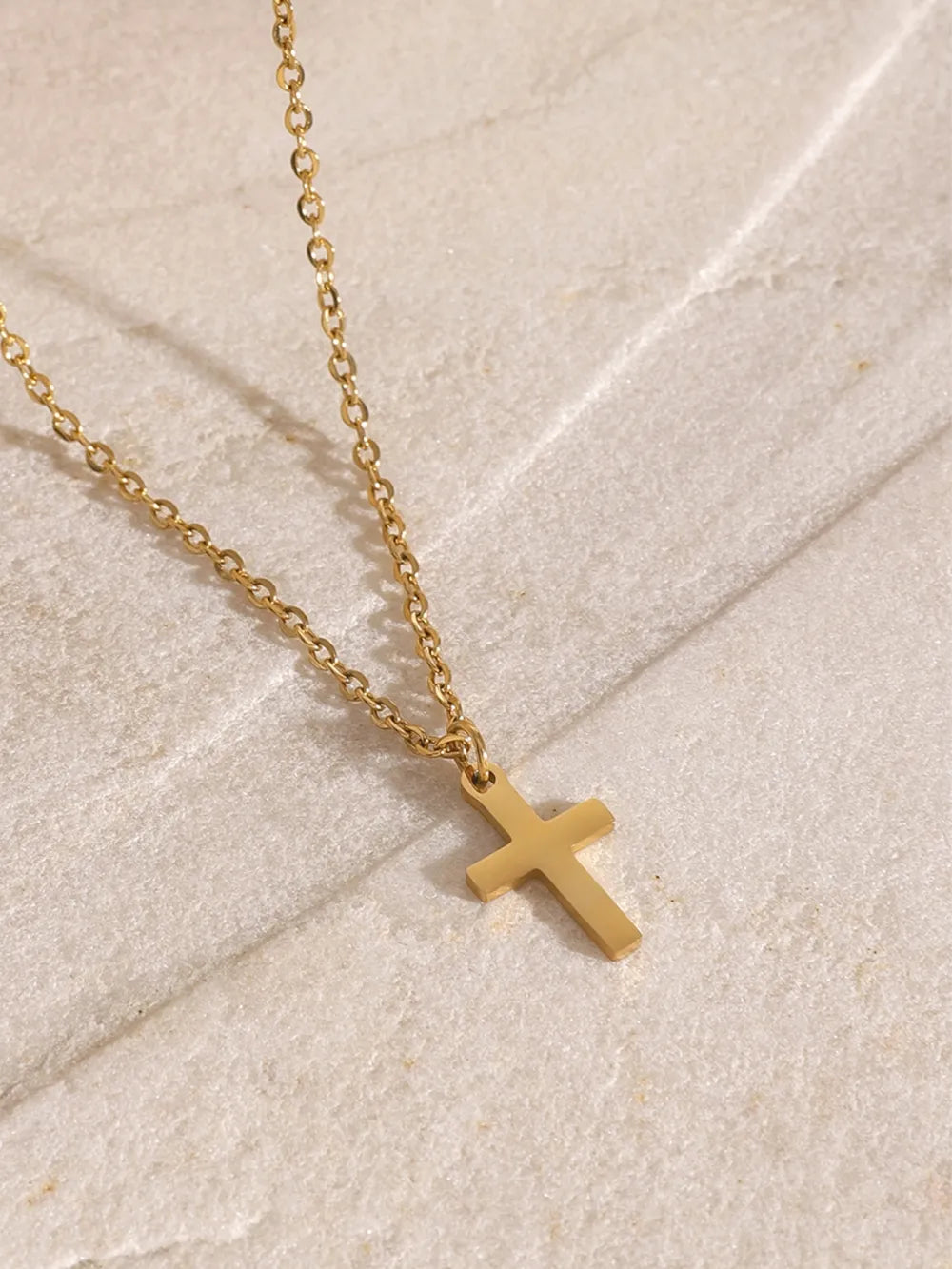 Close-up of a gold-plated cross pendant on a delicate chain, resting on a textured light surface.
