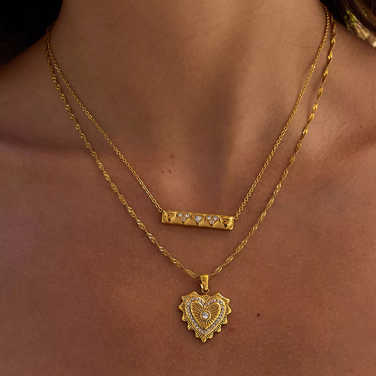 Two layered 18K gold-plated necklaces featuring a zircon-accented heart pendant and a bar pendant on a model's neck.