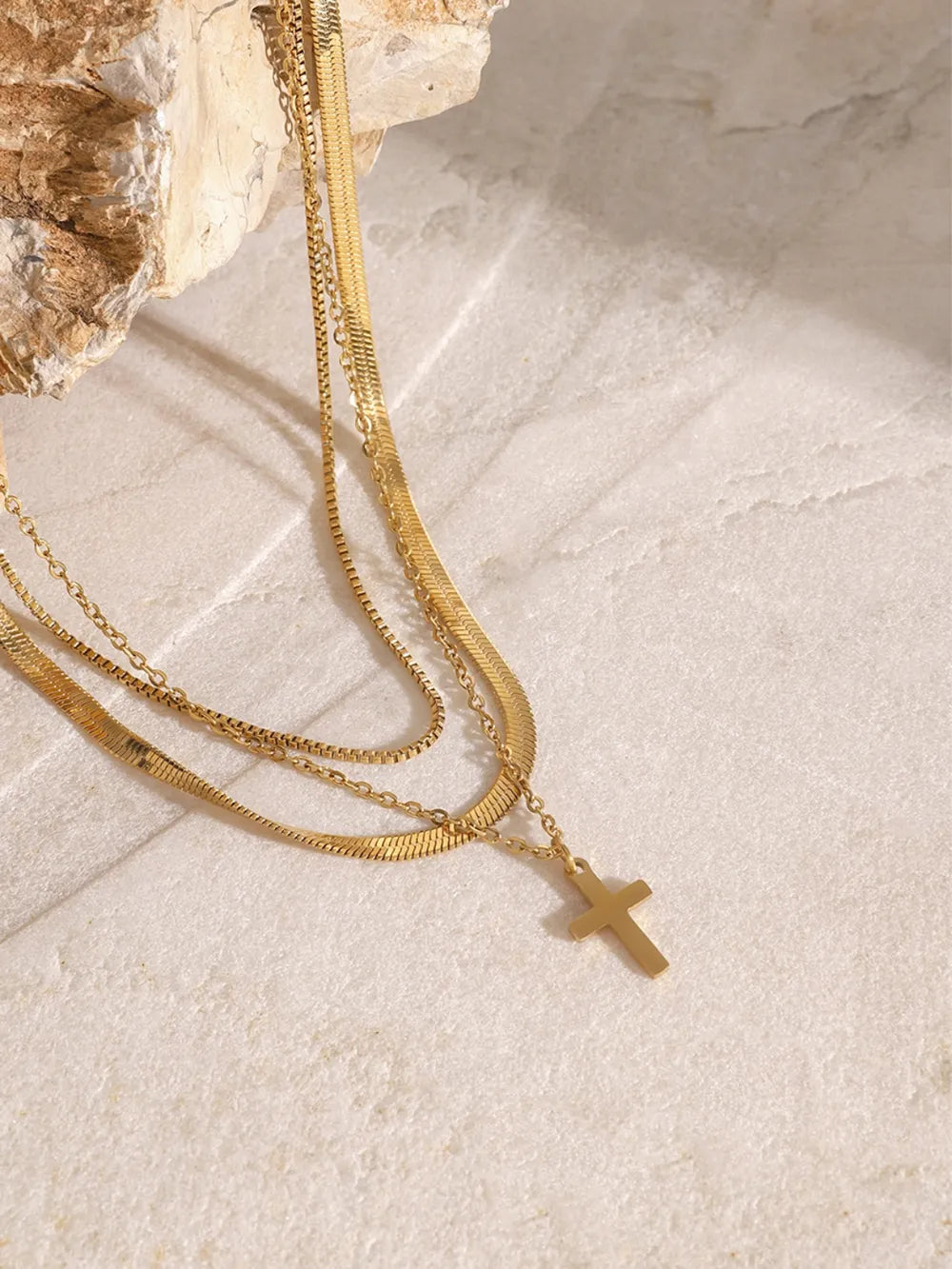 Three-layered 18K gold-plated cross necklace with varied chain styles displayed on a textured stone surface.