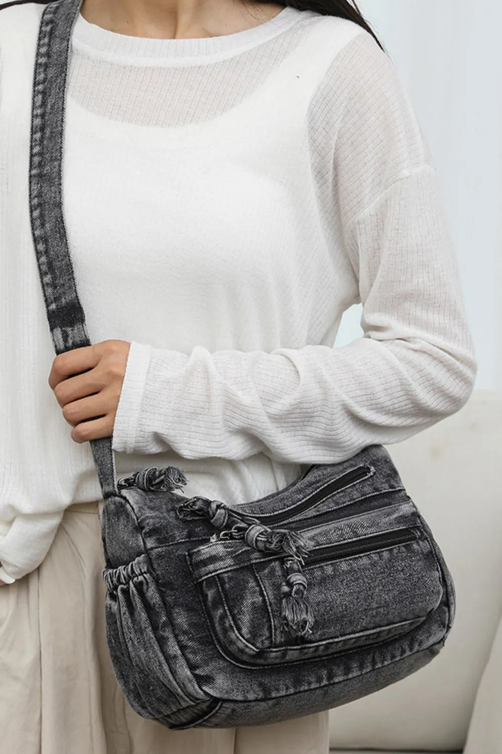 Woman wearing a black denim crossbody bag with multiple zippered pockets and knotted drawstring details