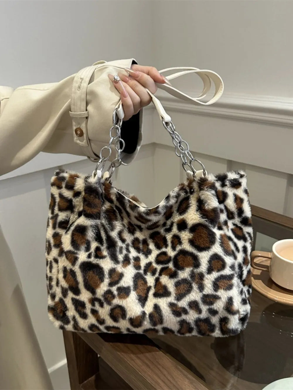 Large leopard print faux fur shoulder bag with chain and leather straps held by a person in a beige coat