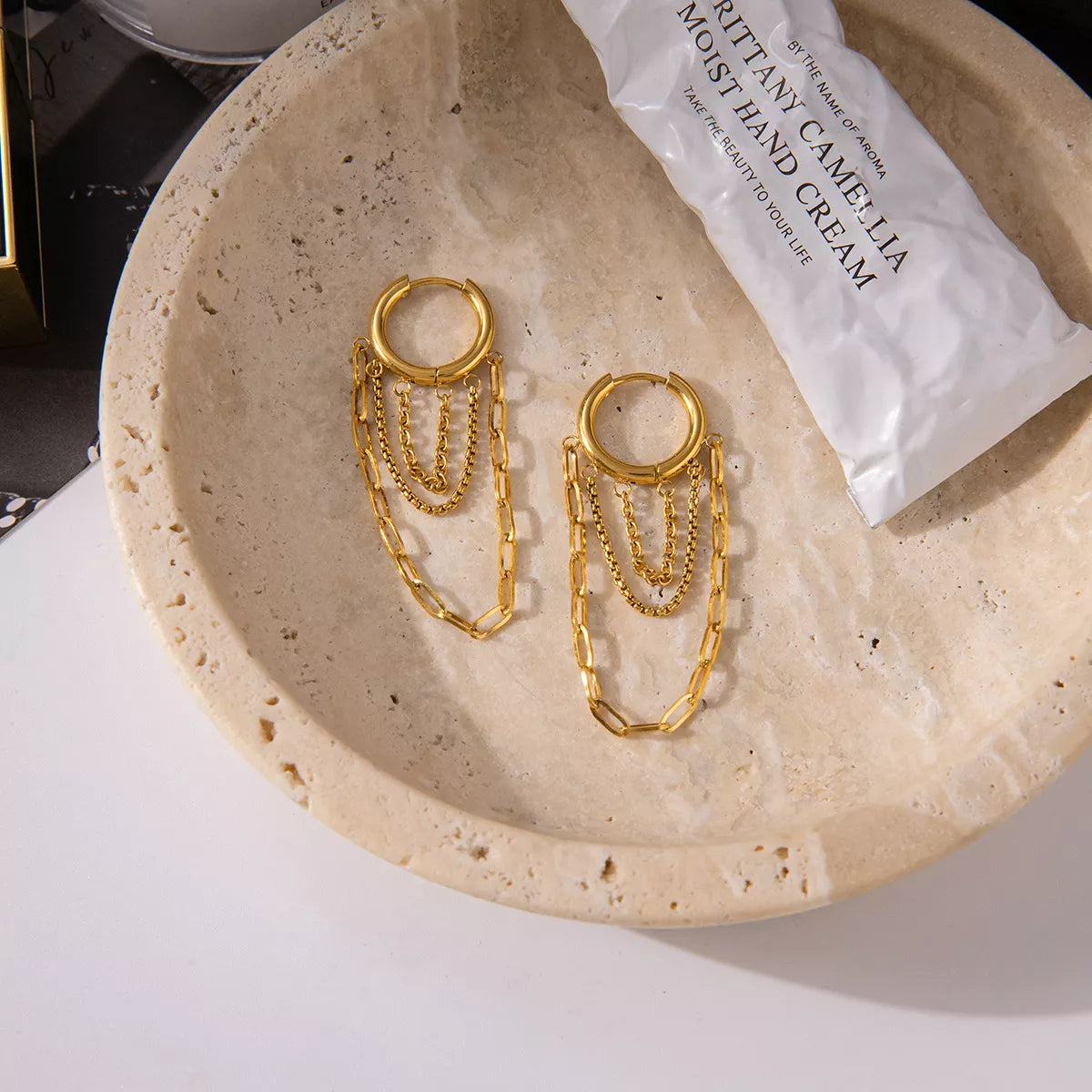 Gold plated chain hoop earrings with multiple delicate chains, displayed on a textured beige dish next to hand cream tube