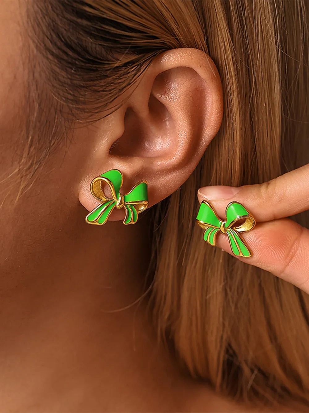 Close-up of green and gold bow stud earrings worn on ear and held between fingers.
