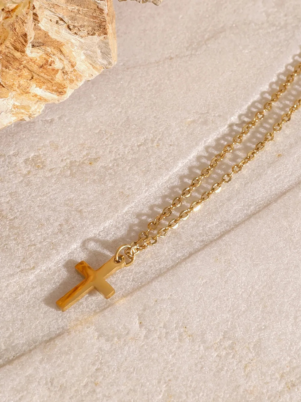 Close-up of an 18K gold-plated cross pendant on a delicate gold chain resting on a textured stone surface.
