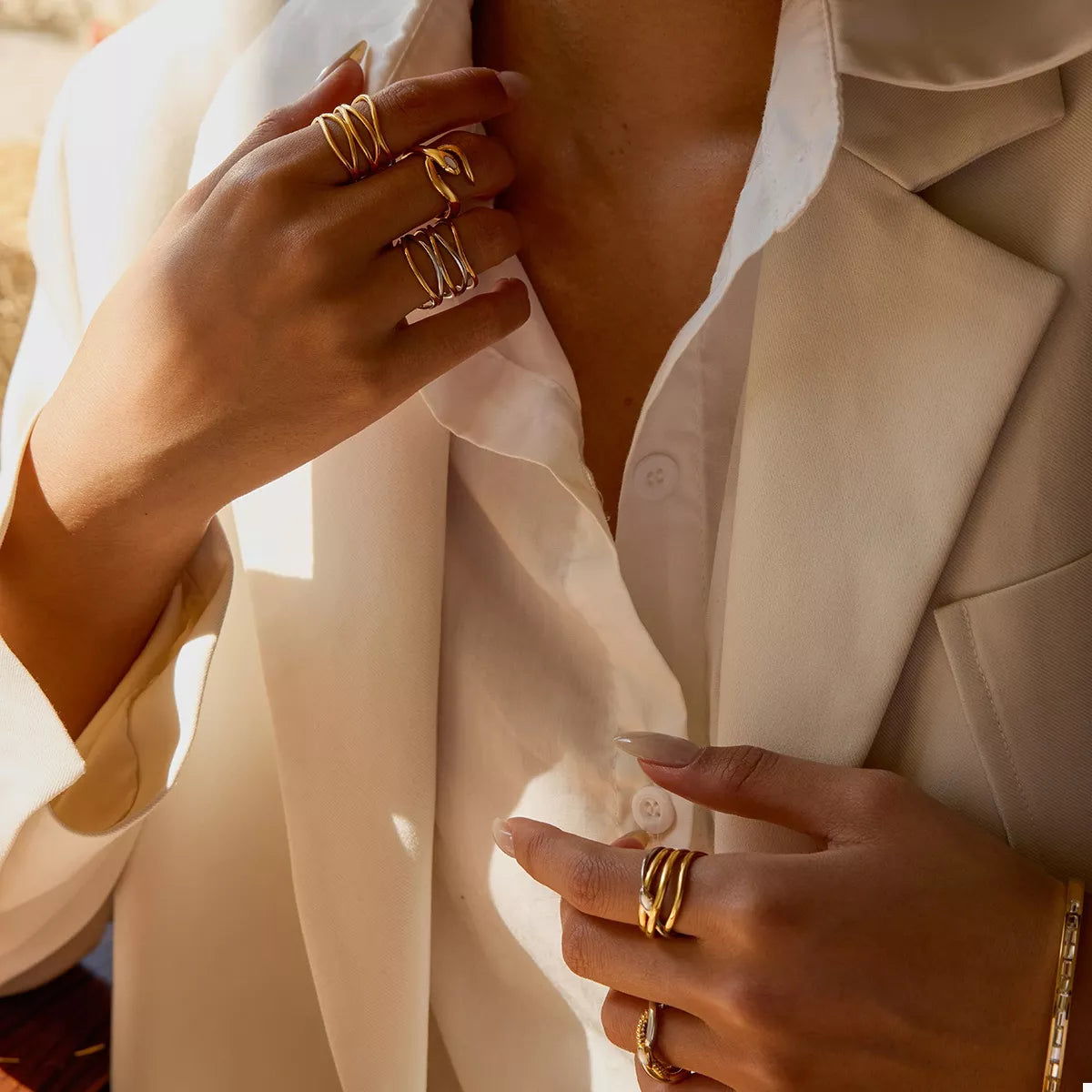 Close-up of a person wearing multiple 18K gold-plated two-tone open rings on fingers with a beige blazer and white shirt.