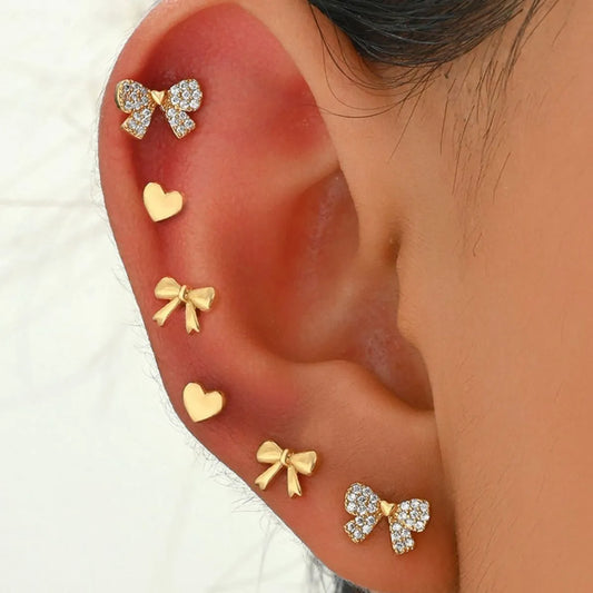 Close-up of an ear wearing gold-plated bow and heart stud earrings with rhinestone accents in a set of six pairs