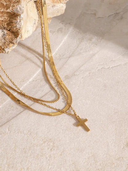 Three-layered 18K gold-plated cross necklace with varied chain styles displayed on a textured stone surface.